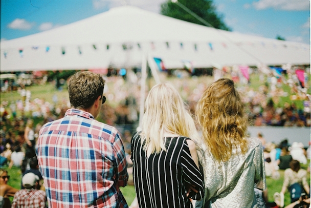 The backs of three people who look out over an outdoor event while the sun shines