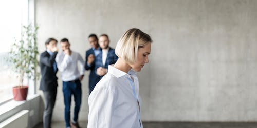 Male Colleagues Whispering Behind Back Of Unhappy Business Lady