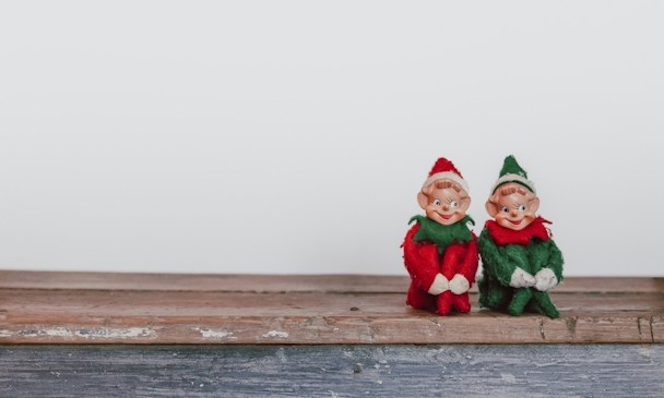 Two Christmas elves sit side-by-side on a shelf