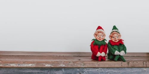 Two Christmas elves sit side-by-side on a shelf