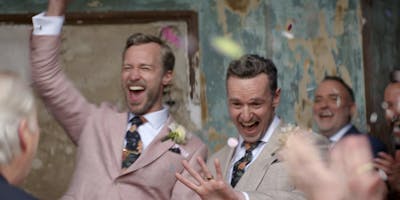 Two men in suits emerging from their wedding ceremony, one with his arm raised in celebration, the other looking at his wedding ring, as guests throw confetti