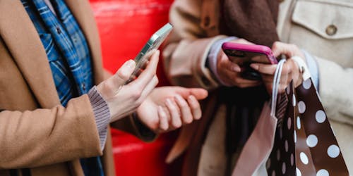 Two women shop on social media on their phones while holding shopping bags